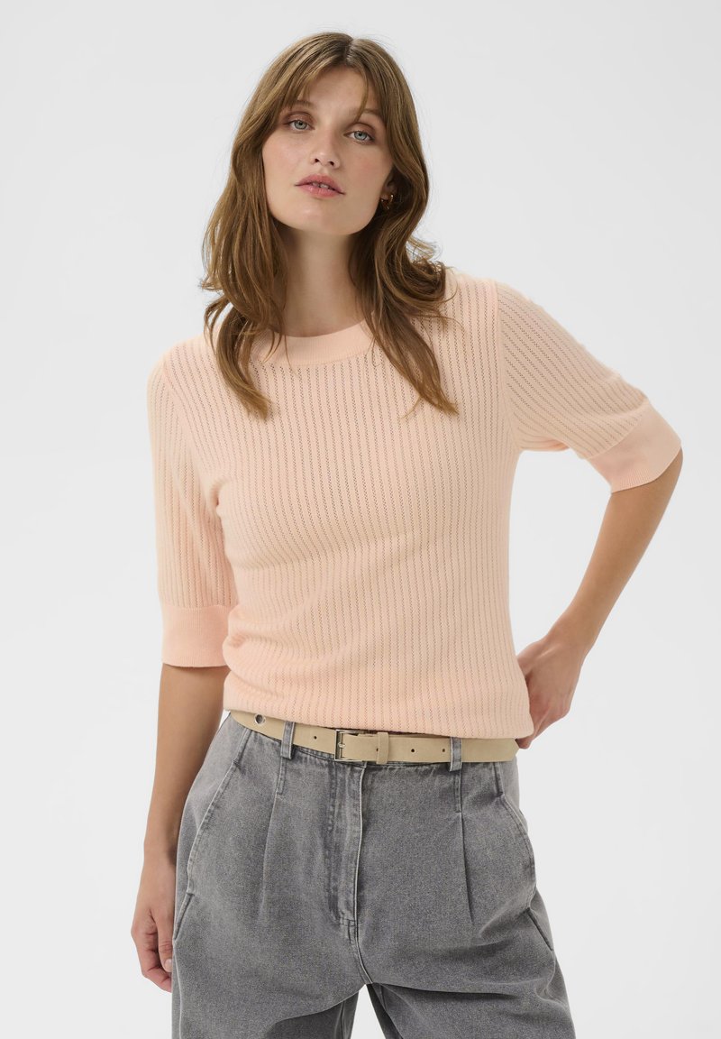 Eine Frau mit hellbraunen Haaren trägt ein blasspfirsichfarbenes Strickoberteil mit Ellenbogenärmeln und einen hellbeigen Gürtel über hochgeschnittenen grauen Jeans, während sie mit einer Hand auf der Hüfte posiert.