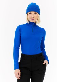 Plavi dugi rukav majica s visokim ovratnikom i zatvaračem sprijeda, uparena s crnim hlačama. Tkanina izgleda glatko, s suptilnim detaljima brendiranja.