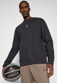 Schwarzer Kapuzenpullover mit einem kleinen weißen Jumpman-Logo, kombiniert mit beigen Jogginghosen und einem schwarzen Logo. Ein strukturiertem Basketball wird in einer Hand gehalten.