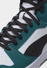 Κοντινό πλάνο ενός λευκού και τιρκουάζ αθλητικού παπουτσιού με μαύρα κορδόνια, που δείχνει λεπτομενές ράψιμο και υφασμένες δερμάτινες επιφάνειες.