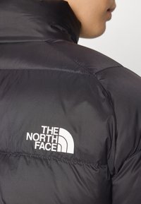 Čierna nafukovacia bunda s hladkou textúrou a prešívanými panelmi. Obsahuje výrazné biele logo "THE NORTH FACE" na ramene.