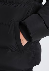 Giacca a vento nera con design trapuntato, dotata di una tasca laterale. Il materiale appare liscio con una finitura lucida, che enfatizza il calore.