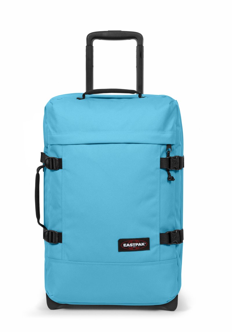 Valigia a rotelle in tessuto azzurro chiaro con manico retrattile, cinghie laterali e frontali, e una patch nera con logo sulla parte anteriore.