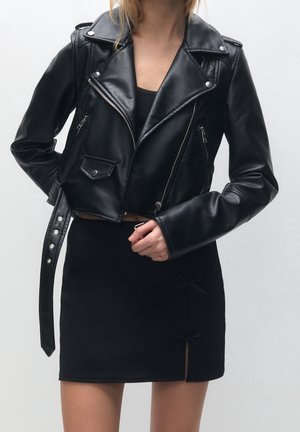 Frau trägt eine schwarze, kurz geschnittene Lederjacke mit Reißverschlusstaschen und einen schwarzen Rock mit zwei kleinen Schleifendetails nahe dem Saum.