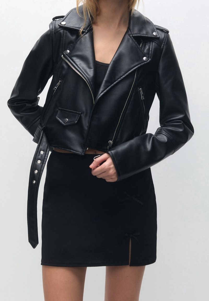 Femme portant une veste en cuir courte noire avec des poches zippées et une jupe noire avec deux petits détails de nœud près de l'ourlet.