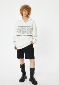 Witte rits-sweater met de tekst "MONTECARLO ATHLETIC VIBES", zwarte strepen; gecombineerd met zwarte shorts en zwarte laarzen tot de knie.