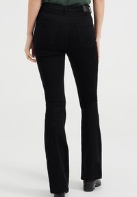 Black flared jeans with a mid-rise waist, made of stretchy denim. Features two back pockets and a label tag on the waistband.