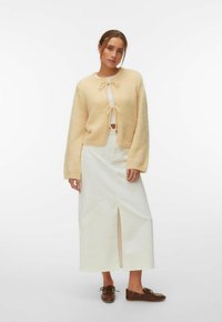 Femme portant un cardigan en tricot crème avec des nœuds papillon, une jupe midi blanche avec une fente avant, un haut blanc et des mocassins en cuir marron, debout sur fond blanc.