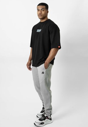 Homme debout, les mains dans les poches, portant un t-shirt noir oversize et un pantalon de jogging gris avec des baskets blanches et noires sur fond blanc.