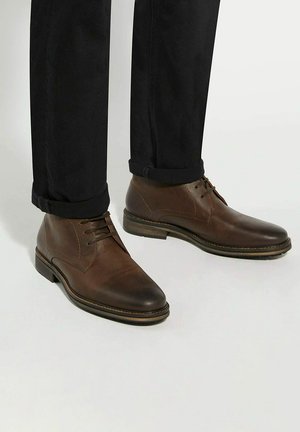 Brown leather ankle boots with a rounded toe, lace-up design, stitched accents, and a low stacked heel, paired with dark trousers.