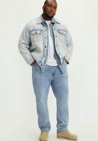 Casaco de ganga de tom mais claro, com dois bolsos no peito, sobre uma camisa azul. Jeans azul claro e sapatos bege completam o visual.