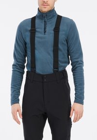 Fleece pullover in teal met een kwart ritssluiting; verstelbare zwarte bretels bevestigd aan zwarte broeken met drukknopen en zijzakken.