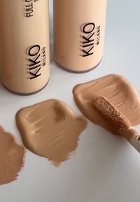 Drei Foundation-Flaschen von KIKO Milano in Beige, begleitet von Mustern der Foundation in verschiedenen Texturen und Nuancen auf einer weißen Oberfläche.