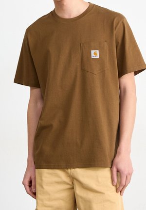 Brązowy T-shirt z krótkim rękawem i okrągłym dekoltem, z kieszenią na piersi z logo Carhartt, noszony z jasnobeżowymi spodniami.