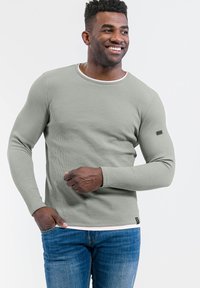 Hellgrünes Langarmshirt mit gerippter Textur, Rundhalsausschnitt und kontrastierendem weißen Besatz, kombiniert mit blauen Jeans.