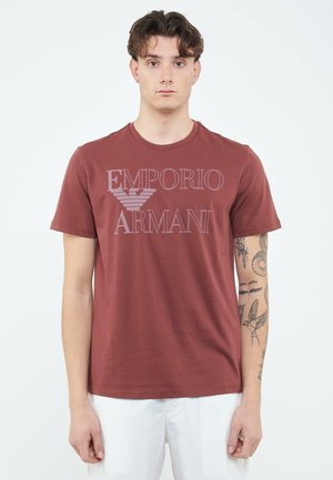 Jonge man met kort donker haar, gekleed in een bordeaux Emporio Armani T-shirt en witte broek, staand tegen een effen witte achtergrond.