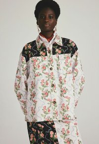 Jas met bloemenpatroon, voorzien van een witte basis met roze en groene rozen, zwarte accenten op de schouders en twee voorzakken.
