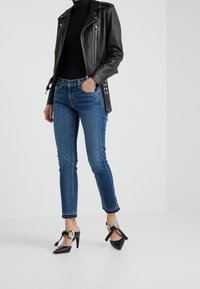 Chaqueta de motorista de cuero negra sobre un jersey de cuello alto negro; jeans ajustados azules con dobladillos deshilachados; zapatos de tacón negros con detalle de lazo.