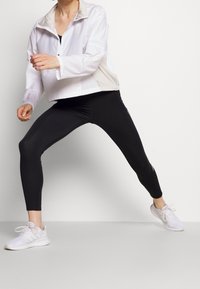 Biała, lekka kurtka zapinana na zamek z neutralnym kołnierzem, w komplecie z czarnymi legginsami. Modelka w dynamicznej pozie, nosi białe buty sportowe.