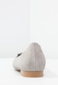 Zapato plano de ante gris claro con suela de goma baja color beige, visto desde atrás sobre una superficie y fondo blancos.