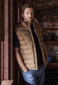 Muž s vousy, oblečený v béžové prošívané vestě a černé košili, stojící uvnitř se založenýma rukama v kapse, opřený o dřevěný rám a kamennou zeď v pozadí.