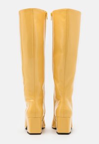 Bottes jaunes montantes en matériau brillant, dotées d'un talon carré et de zips à l'arrière. Texture lisse avec un minimum de détails.