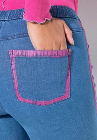 Denim jeans met een unieke blauwe kleur en een roze accent rond de achterzak, met een textuurafwerking en een metallic knoopdetail.