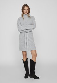 Robe en tricot gris avec un col montant, une ceinture à nouer et un ourlet effiloché, associée à des bottes noires en daim arrivant au genou avec un talon bas.