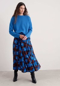 Donna che indossa un vivace maglione blu e una gonna plissettata marrone con grandi motivi floreali blu, abbinata a stivali neri fino al polpaccio, in piedi all'interno.