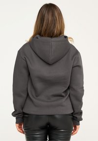 Kapuzensweatshirt in Dunkelgrau mit bequemer Passform, gerippten Säumen und einem strukturierten Stoff. Getragen mit schwarzen, glänzenden Hosen.