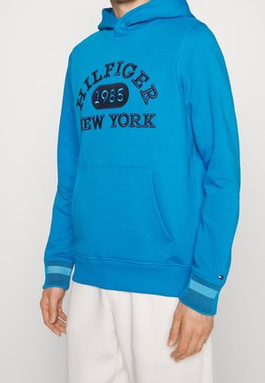 Sweat à capuche bleu en coton, avec une poche avant, des poignets côtelés et un logo brodé "HILFIGER 1985 New York" en noir.