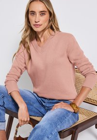 Un pull-over rose clair en V avec des poignets et un ourlet côtelés, porté avec un jean bleu, assis sur une chaise avec des détails de siège en osier.