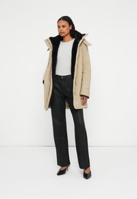Beige parka met zwarte bontvoering en ritssluiting, gedragen over een grijze top en zwarte wijde broek. Zwarte hoge hakken.