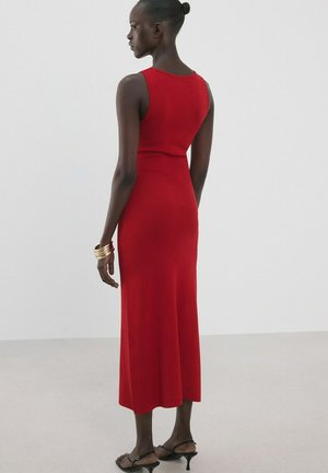 Femme à la peau foncée et aux cheveux attachés en un chignon, portant une robe midi rouge sans manches, des sandales noires à talons et des bracelets dorés, debout devant un fond uni.