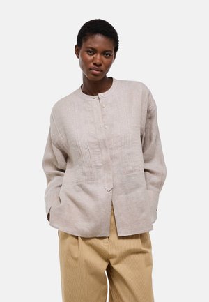 Femme aux cheveux courts portant une chemise texturée beige clair à manches longues et un pantalon en velours côtelé couleur moutarde sur un fond uni.