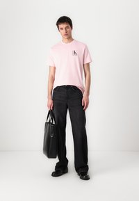 T-shirt rose pâle à manches courtes avec un logo, associé à un jean noir à pattes larges et un sac fourre-tout noir, porté avec des chaussures noires.