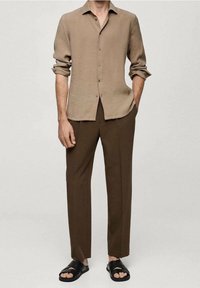 Vaalean beige nappipaita, jossa on pitkät hihat, kevyt kangas ja terävä kaulus, yhdistettynä ruskeisiin slim fit -housuihin ja mustiin liukujalkineisiin.