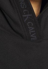 Sudadera negra con un collar alto y el logo de Calvin Klein a lo largo del escote. La tela es lisa, con una textura suave.