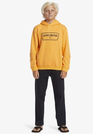 Gelber Hoodie mit schwarzem Logo, Fronttasche, aus Baumwollmischung. Kombiniert mit schwarzen Hosen und Flip-Flops, stehend auf einem schlichten weißen Hintergrund.