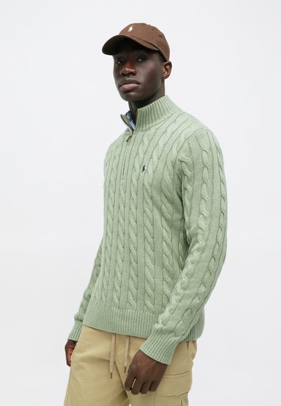 Ανοιχτό πράσινο πουλόβερ με πλέξη καλωδίου και υψηλό κολάρο με φερμουάρ. Καφέ καπέλο μπέιζμπολ με λογότυπο. Απαλό μπλε κολάρο πουκαμίσου ορατό από κάτω.