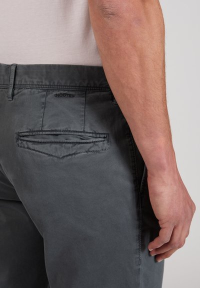 Primo piano del braccio destro di una persona accanto a un paio di pantaloni grigi con tasca a filetto posteriore e dettagli di cuciture visibili.
