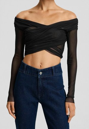 Olkapäät paljastava musta verkkokangas crop-toppi, jossa pitkät hihat, yhdistetty tummansinisiin korkeavyötäröisiin farkkuihin seisovalla henkilöllä.