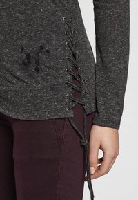 Gray long-sleeve top featuring a lace-up side detail with metallic eyelets and a textured finish, paired with maroon pants.