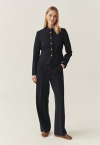 Woman standing in navy tailored blazer with gold buttons and matching wide-leg trousers, wearing brown suede sneakers.