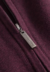 Bordeaux stof met een ribbeltextuur, voorzien van een metalen rits met een label "FRIEDA & FREDDIES." Close-up detail van het ritsgebied.