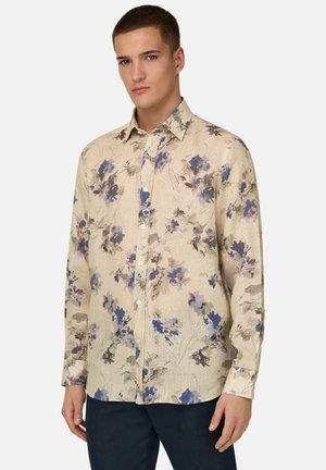 Mand iført beige skjorte med blå og brune blomster, stående foran en ensartet lys baggrund.