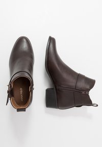 Botas de cuero marrón hasta el tobillo con tacón bajo de bloque, punta puntiaguda, cremallera lateral y acabado liso. Etiqueta de la marca visible en el interior.