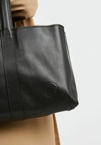 Sac fourre-tout en cuir noir avec une texture lisse, arborant un logo en relief circulaire. Le sac est doté de poignées robustes et d'un design simple.
