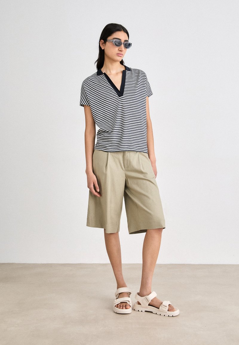 Pasiasty czarno-biały krótki rękaw top z dekoltem w kształcie litery V, zestawiony z khaki szortami do kolan i białymi sandałami z regulowanymi paskami.