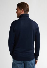 Donkerblauwe gebreide trui met hoge kraag, getailleerd ontwerp, lange mouwen en ribbels aan de onderkant. Gladde textuur met minimale details.